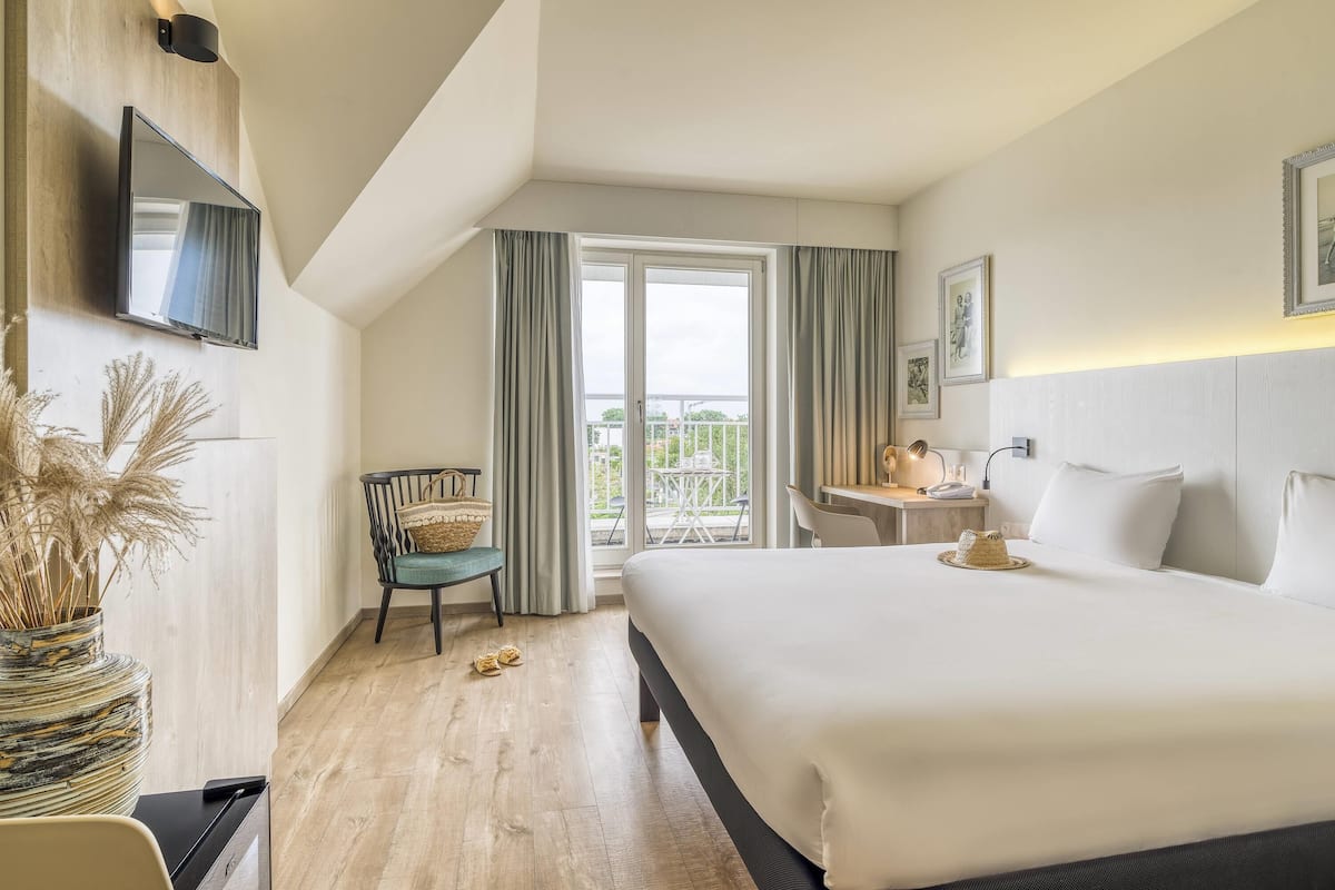 A hotel room with a bed, a desk, a chair, a TV mounted on the wall, and a view of the outside balcony.