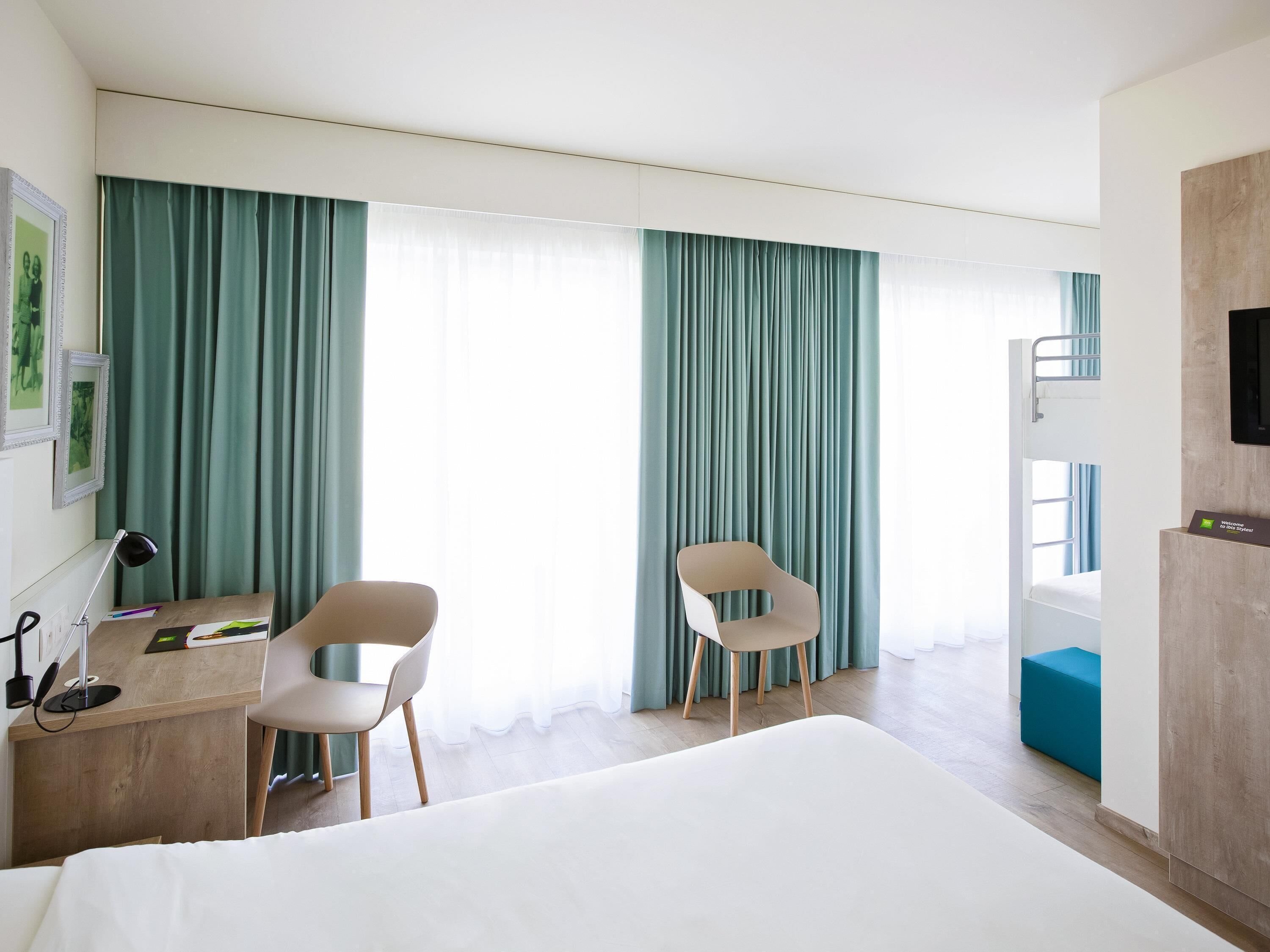 A hotel room with a bed, two chairs, a desk, a TV, and curtains.