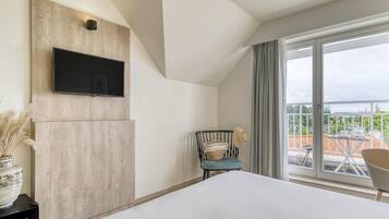 Ein modernes Hotelzimmer mit einem großen Bett, einem Wand-Fernseher, Blick auf die Stadt durch große Fenster und einem Balkon mit Sitzgelegenheit.
