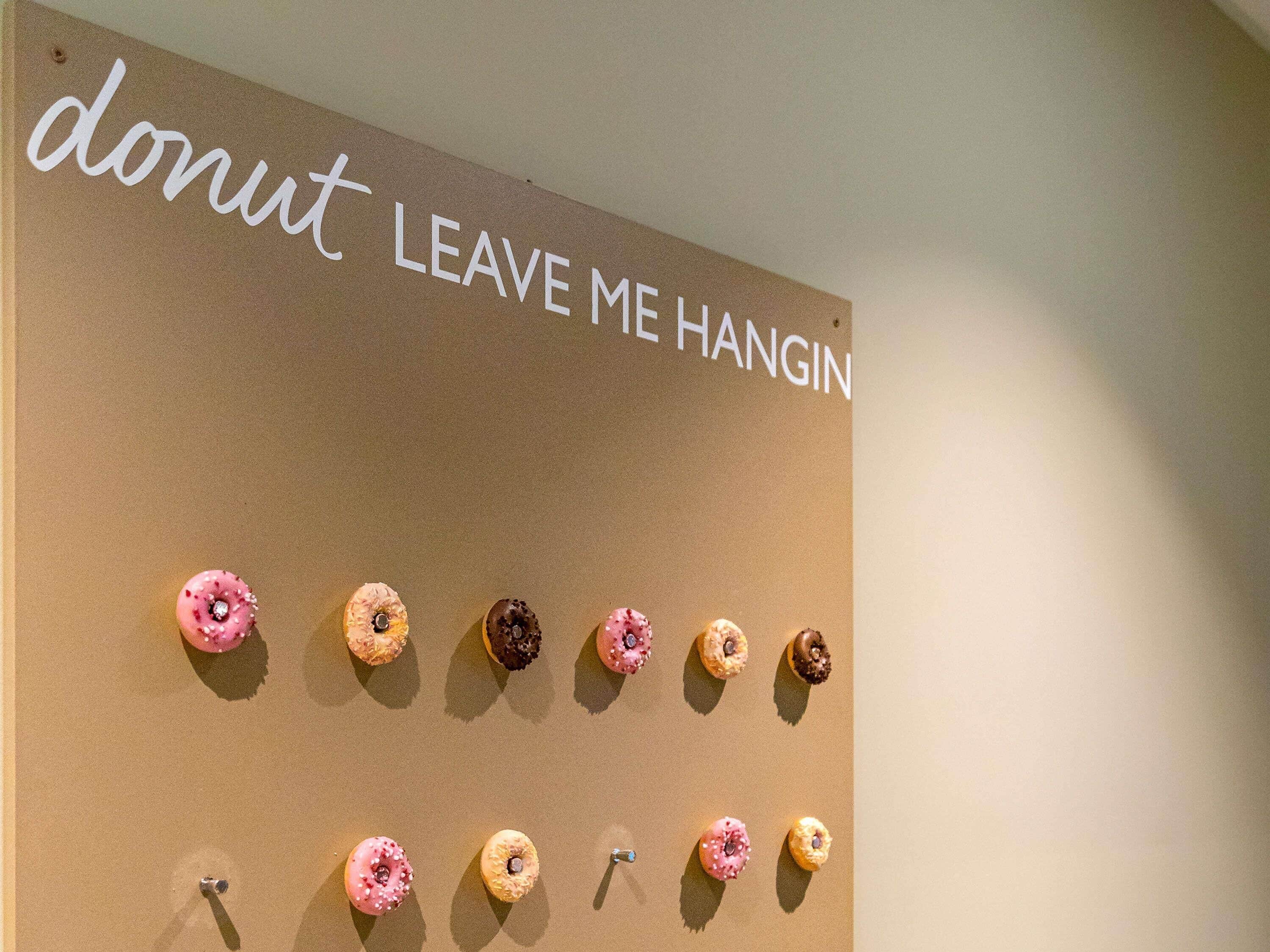 A wall with a sign that reads "donut LEAVE ME HANGIN" and a row of donuts displayed below.