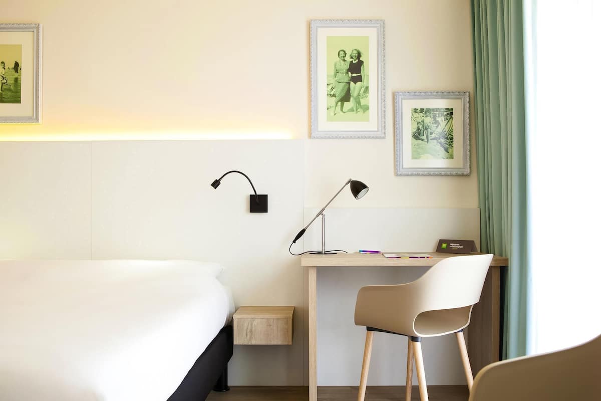 Ein Hotelzimmer mit Bett, Schreibtisch, Stuhl, zwei Wandlampen und gerahmten Bildern.