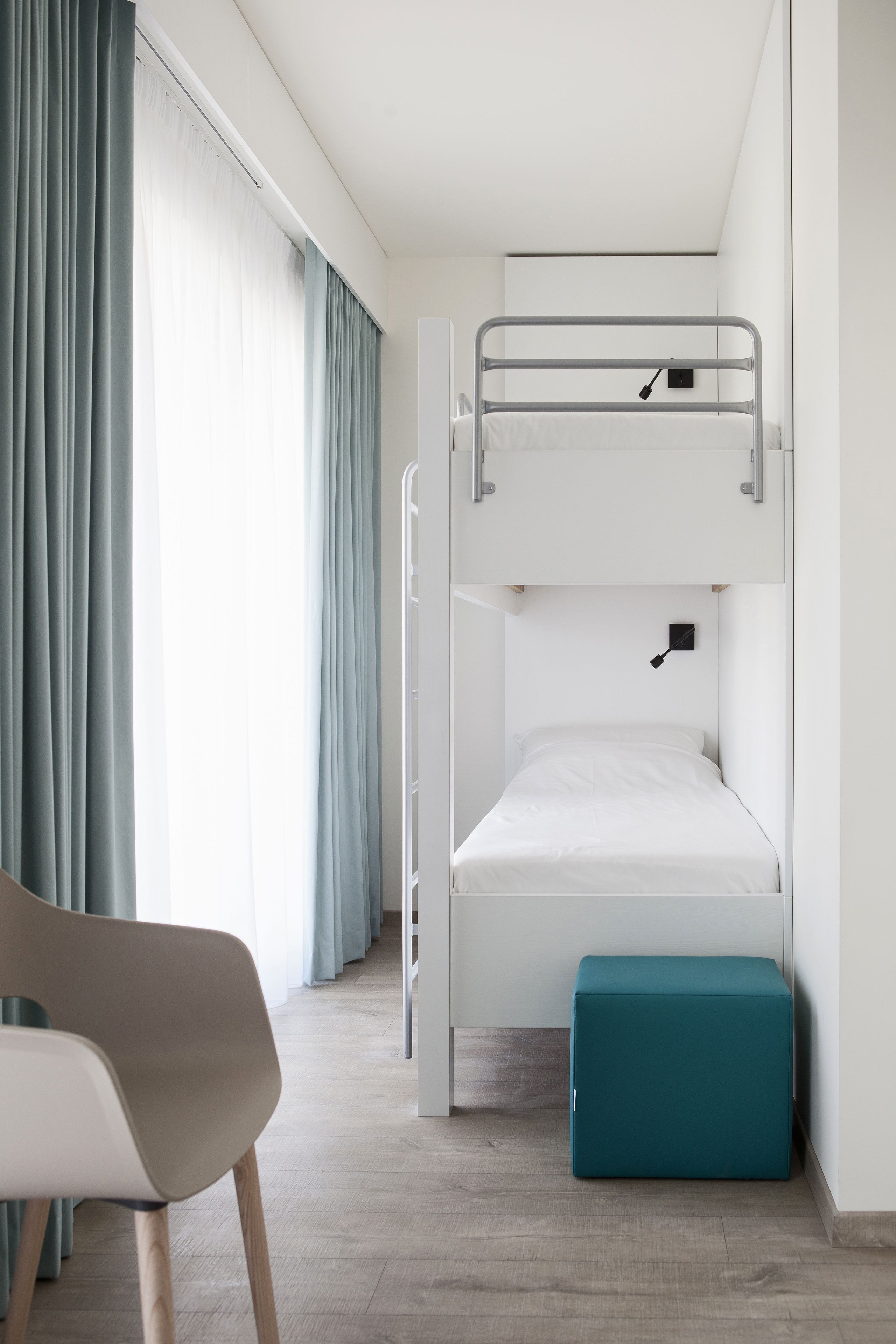 Un lit superposé avec un bureau et une chaise, un pouf turquoise et une fenêtre avec des rideaux.