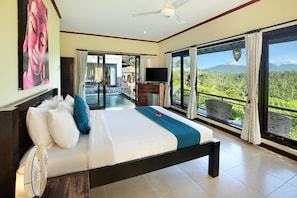Un dormitorio espacioso con una cama grande, televisor y vistas a un paisaje exuberante a través de amplias ventanas.
