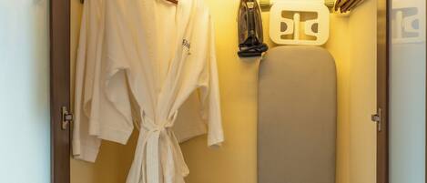 A compact hotel room with a built-in wardrobe, an ironing board, and a small shelf with a control panel.