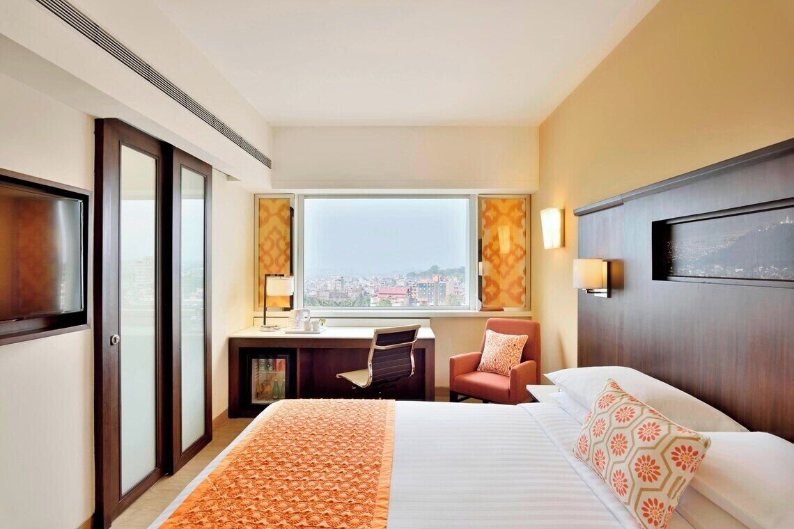 Habitación de hotel con cama, escritorio, silla y un amplio ventanal con vista a la ciudad.