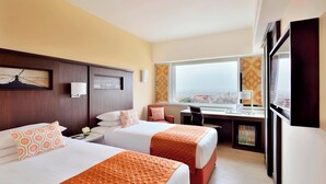 A hotel room with two beds, a desk, a chair, a small fridge, and a large window with a city view.