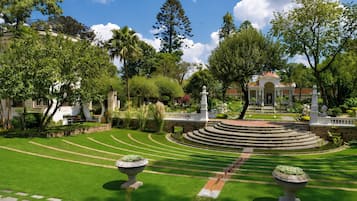 Un jardín cuidado con un estanque circular central, rodeado de varios árboles y un edificio clásico al fondo.