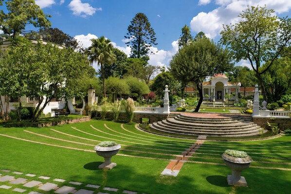 A well-manicured garden with a central circular fountain, surrounded by a variety of trees and a classical building in the background.