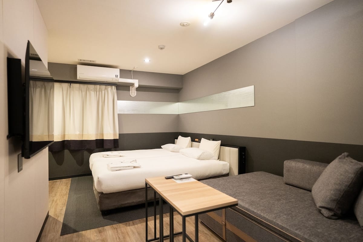 A modern hotel room with a bed, a sofa, a small table, and a mounted air conditioner.