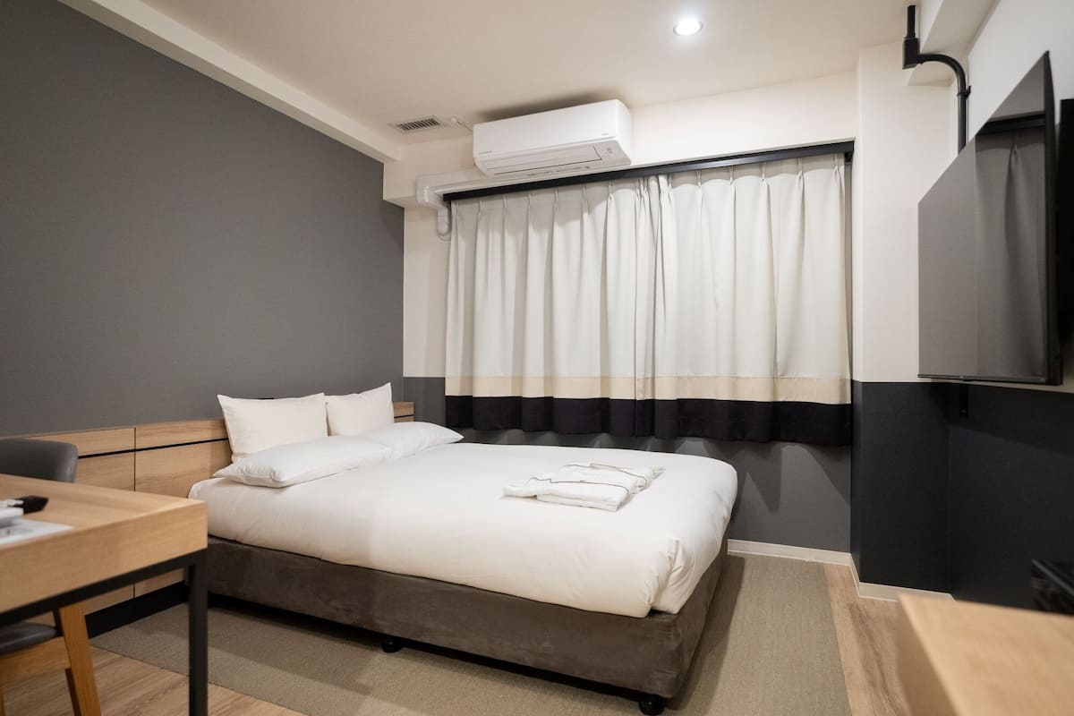 A hotel room with a bed, a desk, a TV, and an air conditioning unit.
