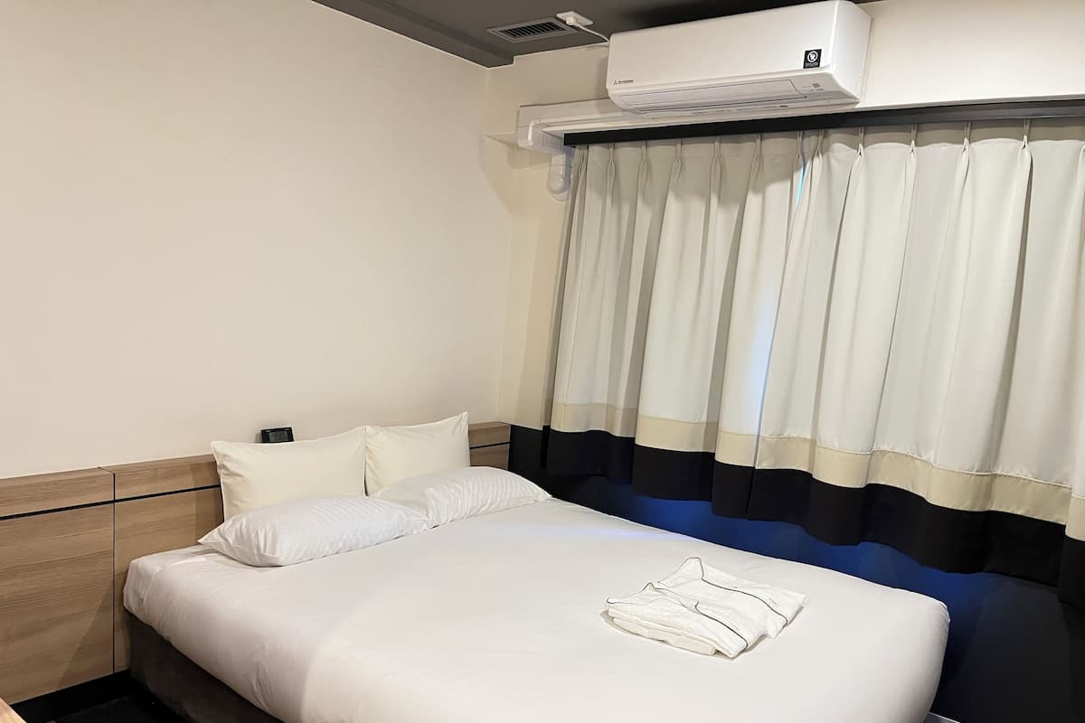 A hotel room with a bed, white bedding, a folded towel, a nightstand, a wall-mounted air conditioner, and a window with curtains.