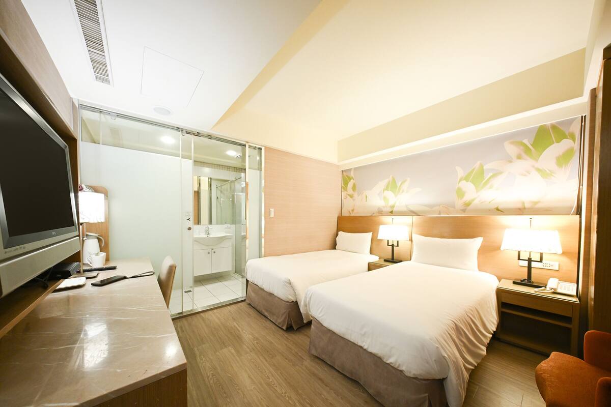 A hotel room with a television, desk, two beds, a bathroom, and a floral wall mural.