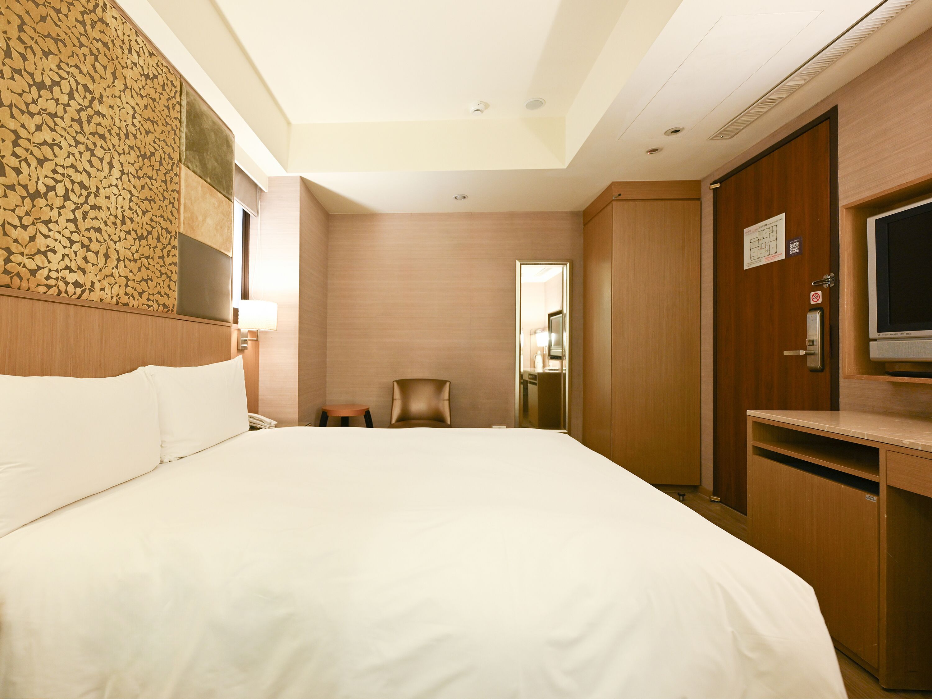 A hotel room with a large bed, a desk with a TV, a chair, and a mirror.