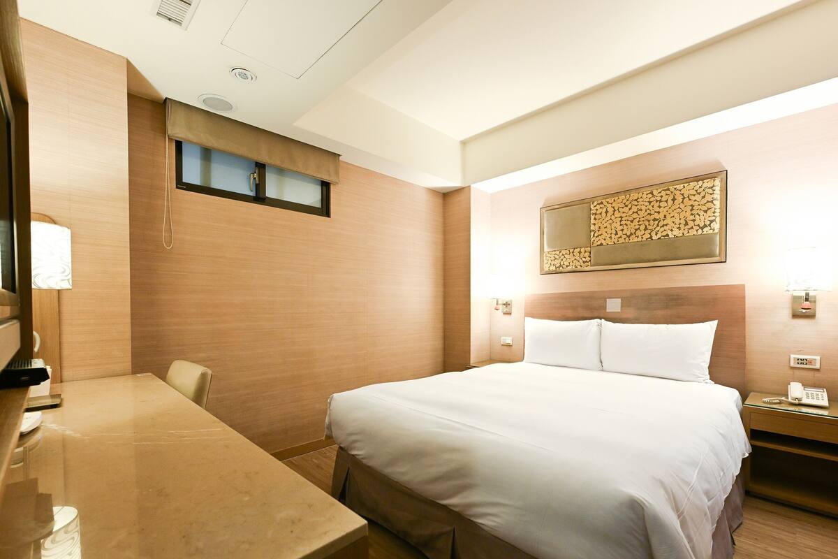 A hotel room with a bed, bedside table, and a desk with a chair.