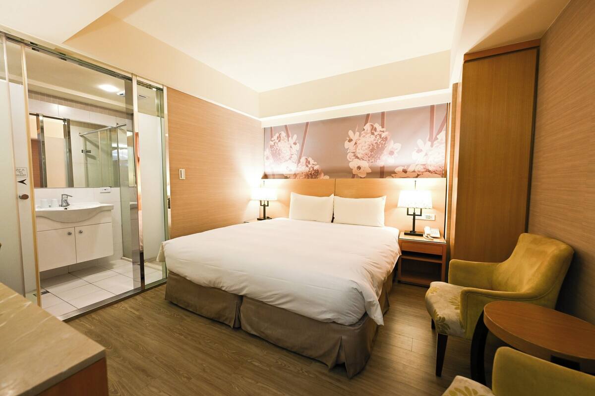 A hotel room with a bed, a chair, a small table, a nightstand, a lamp, a mirror, and a bathroom with a sink and glass door.