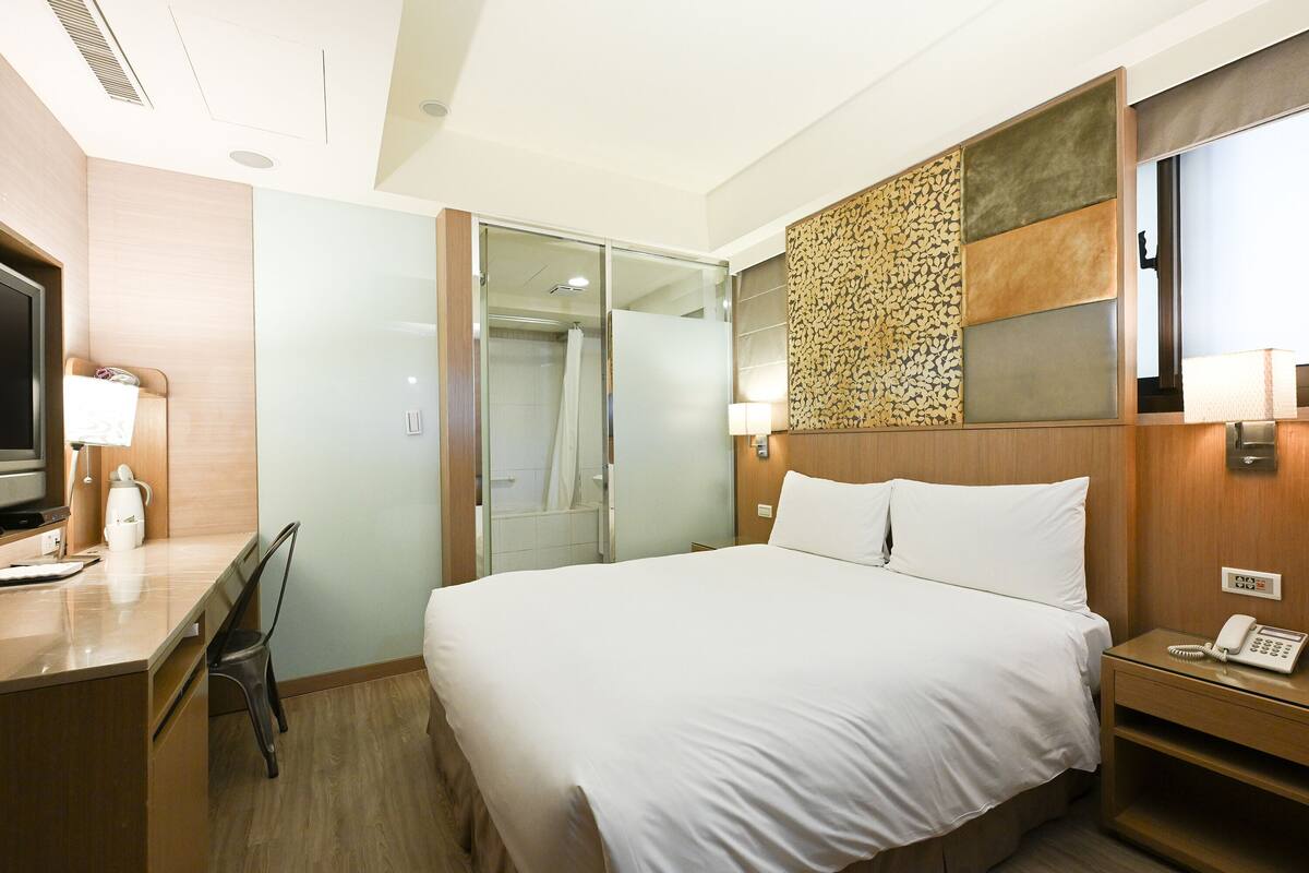 A hotel room with a bed, a desk with a TV, a shower, and a phone on the nightstand.