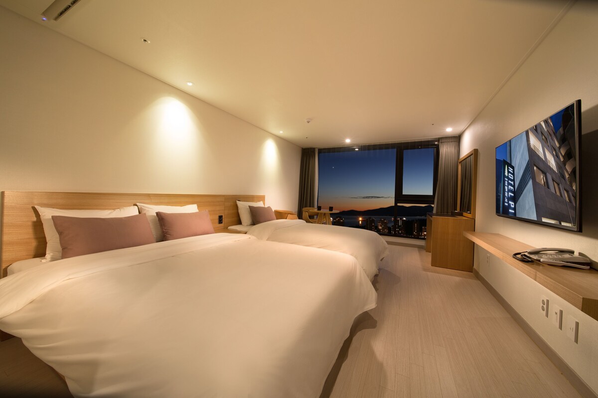 A hotel room with two beds, a large window with a city view, a desk with a phone, and a TV mounted on the wall.