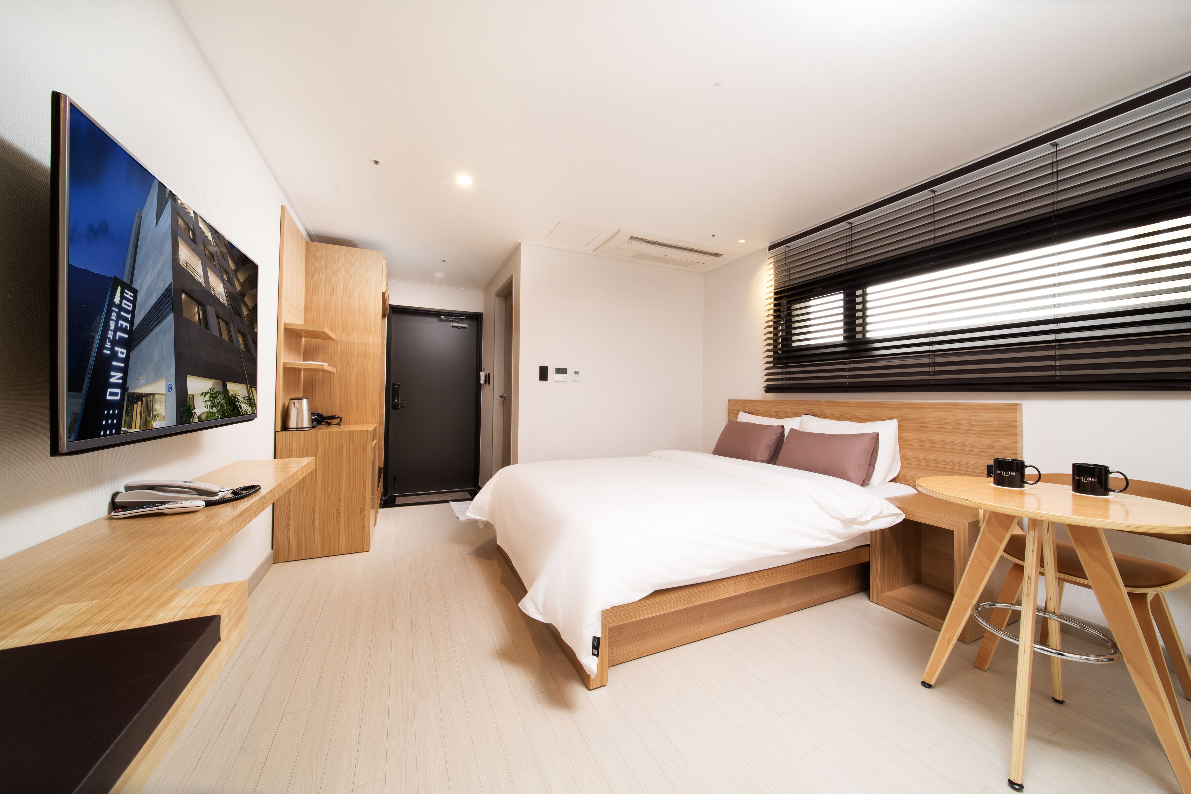A modern hotel room with a bed, a desk, a chair, a TV, and a window with blinds.