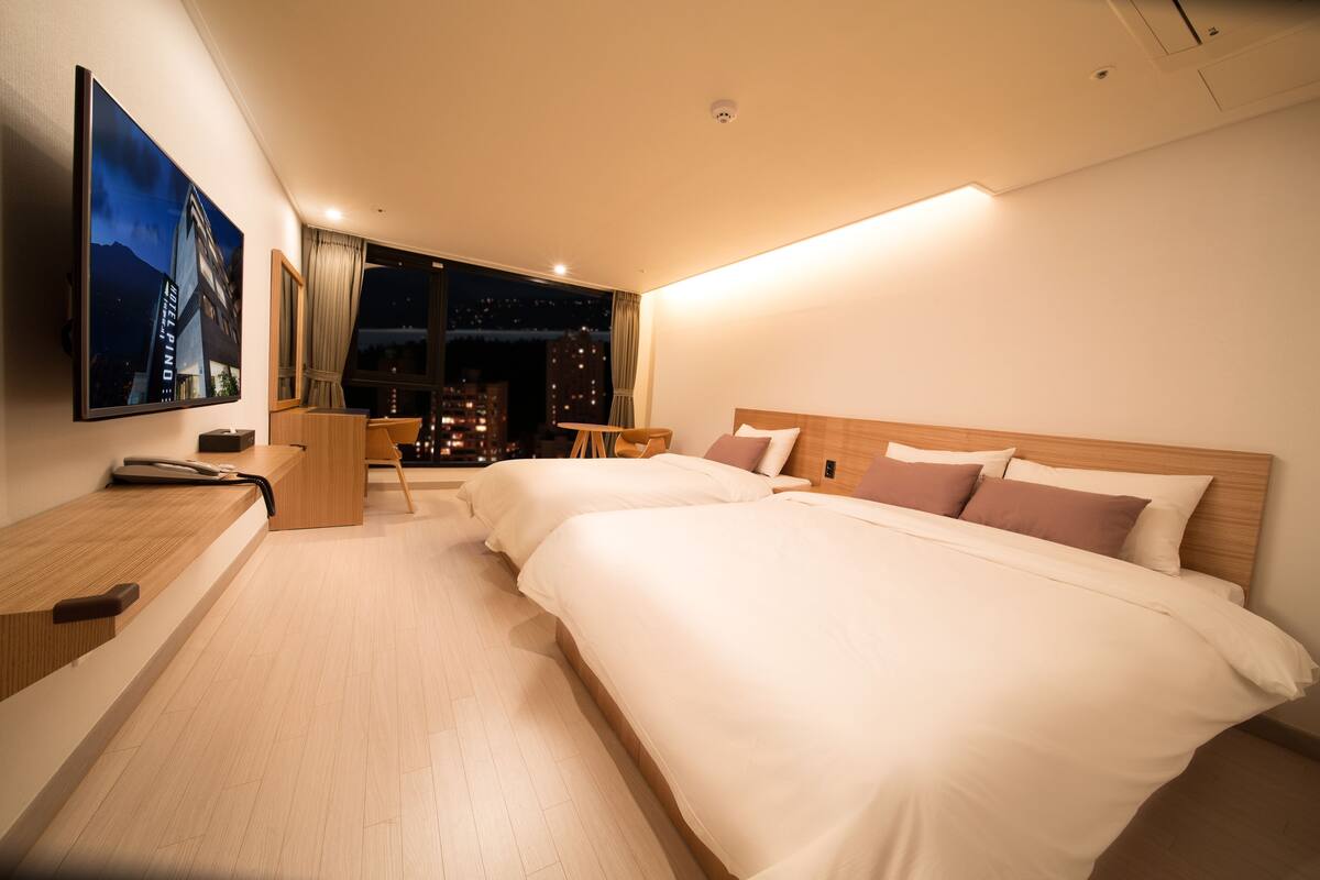 A modern hotel room with two beds, a desk, and a large window offering a city view.