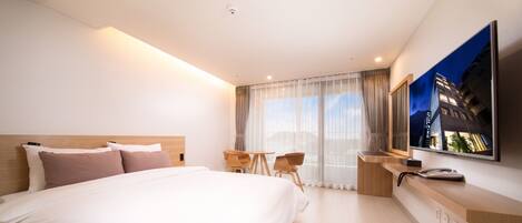 A modern hotel room with a large bed, a desk with a telephone, a chair, and a large window with curtains.