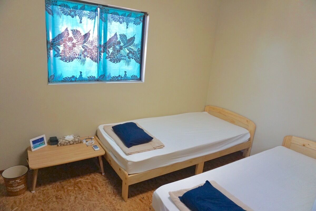 Een kleine kamer met twee houten bedden, een raam met bloemeninrichting, een klein tafeltje met een laptop en een prullenbak.
