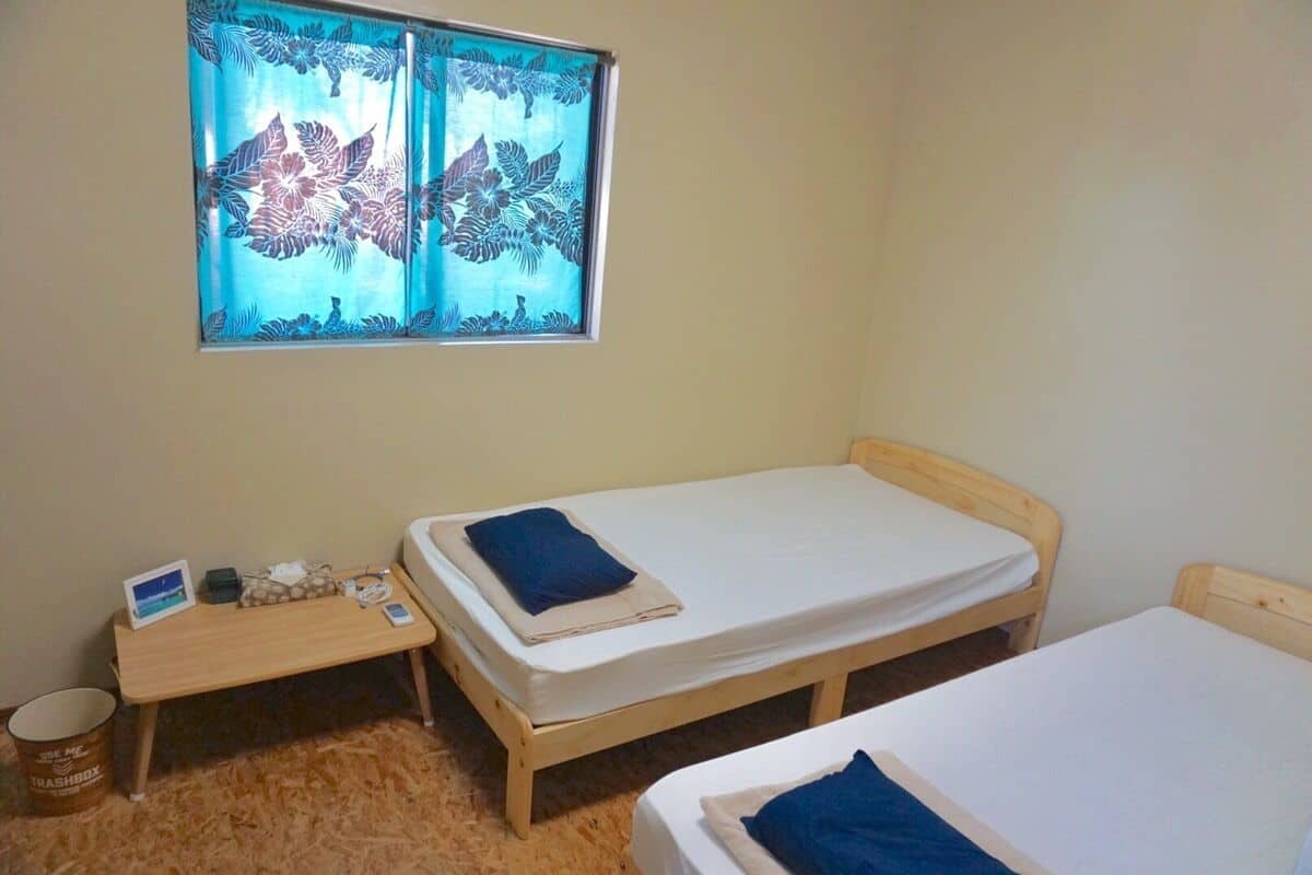 A small room with two wooden beds, a window with a floral curtain, a small table with a laptop, and a trash bin.