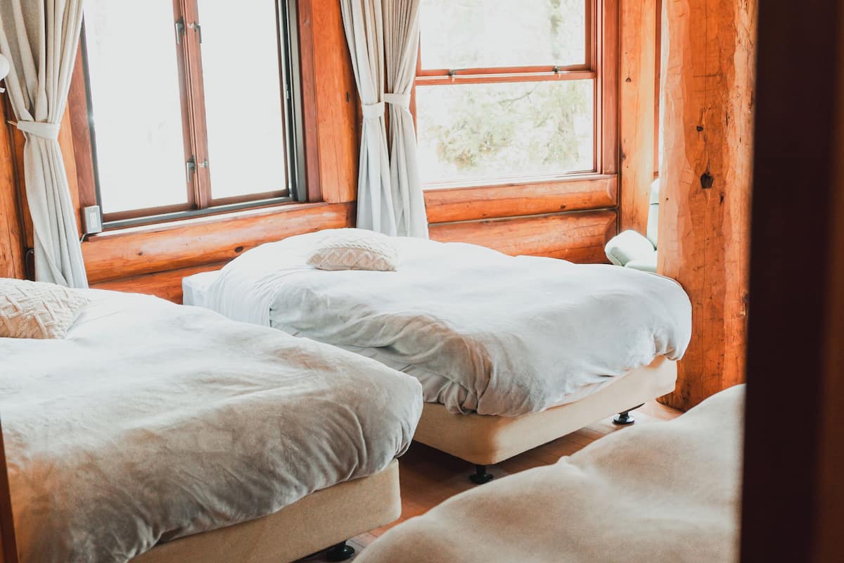 A cozy room with two beds, wooden walls, and large windows with curtains.