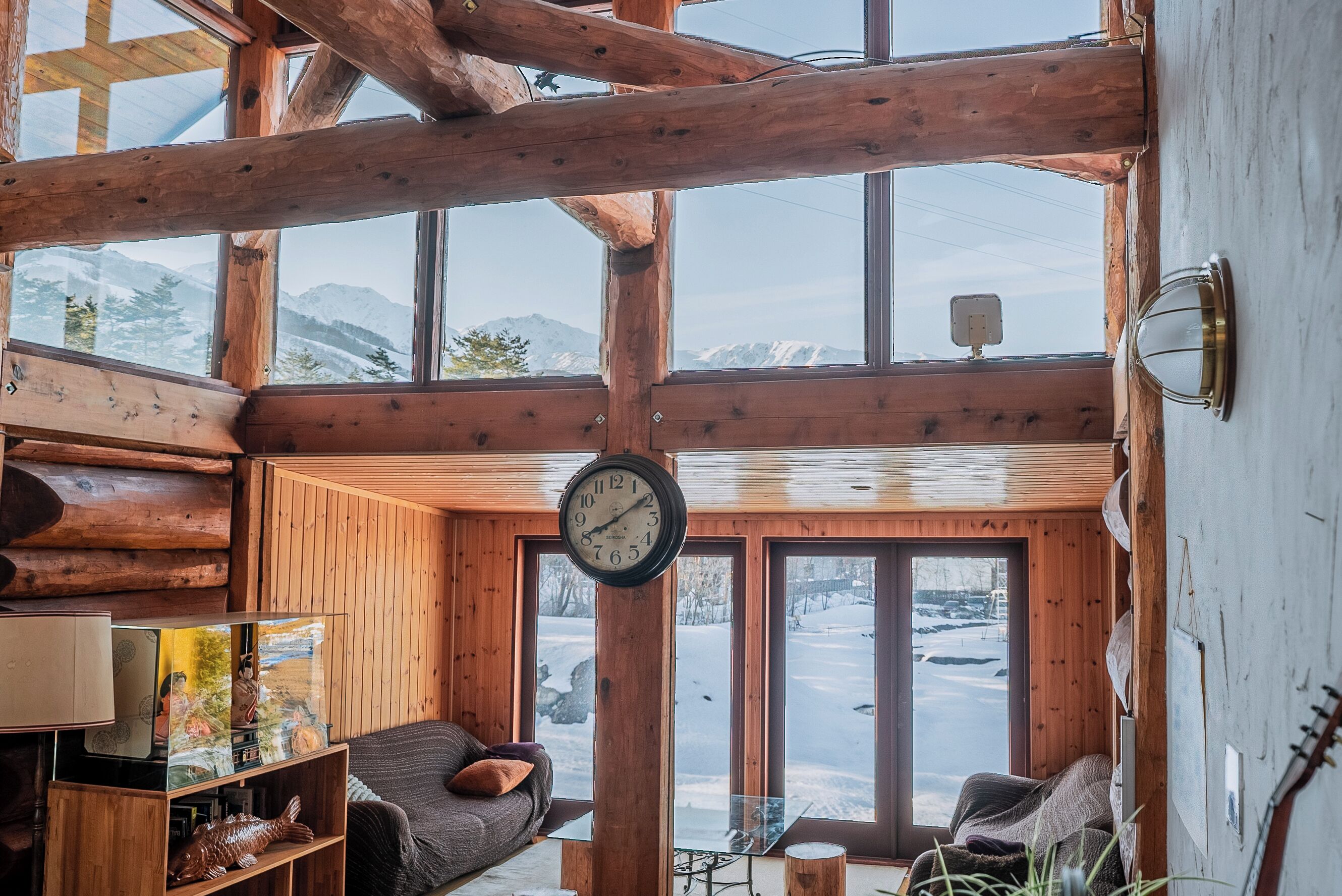 A cozy cabin with a wooden interior, a large window offering a snowy mountain view, a clock on the wall, and a small table with a lamp.
