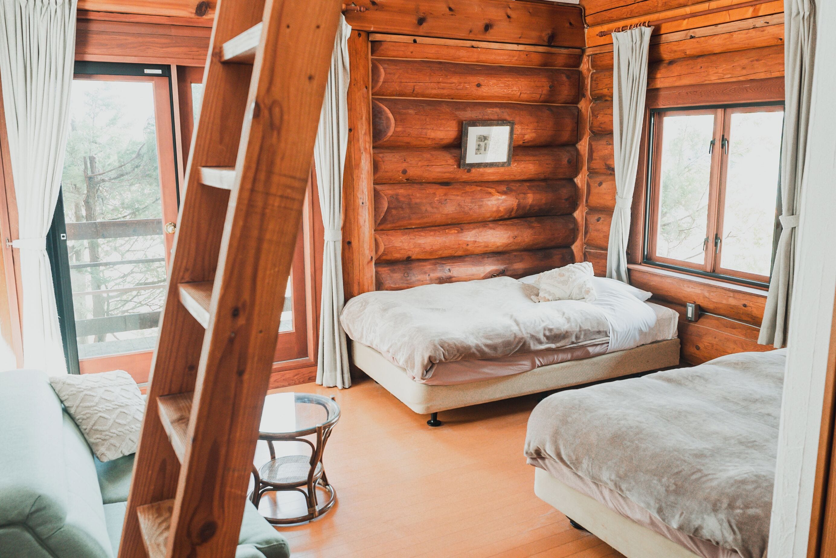 A log cabin bedroom with two beds, a ladder, a small table, and a window with curtains.