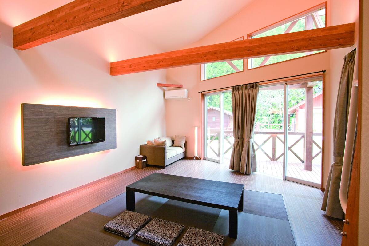 Ein modernes Wohnzimmer mit einer Couch, einem Flachbildfernseher, einer Decke aus Holzbalken und einer Glastür, die auf eine Terrasse im Freien führt.