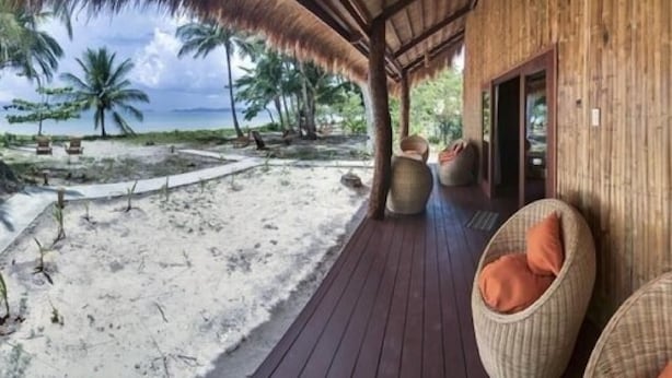 A wooden deck with wicker chairs and cushions, overlooking a sandy beach with palm trees and a clear sky. - QI Palawan by Cocotel powered by ASTON (El Nido)