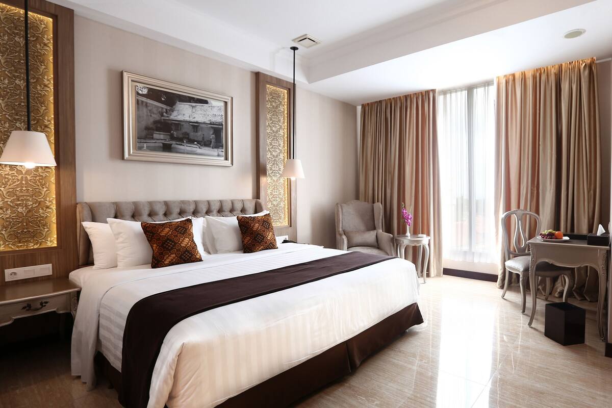 A hotel room with a large bed, a desk, a chair, a small table, and a window with curtains.