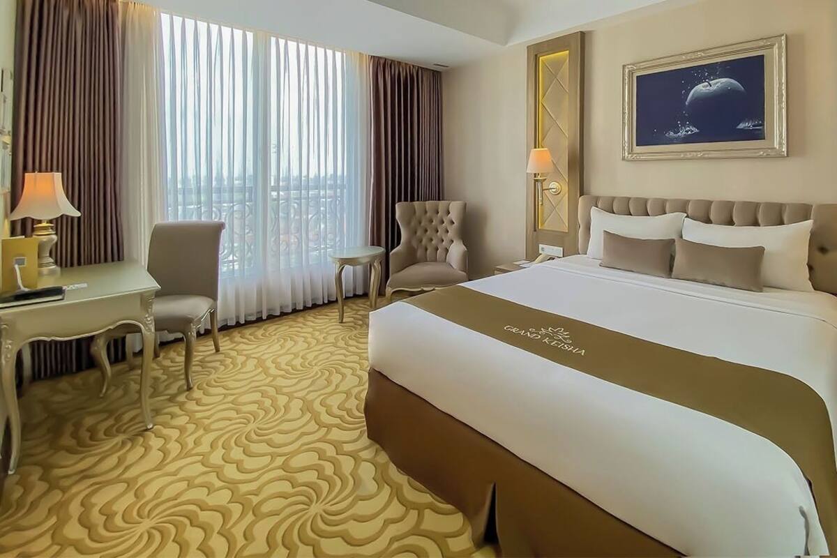 A hotel room with a large bed, a desk, a chair, and a view of the city through large windows.