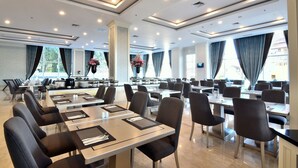 A spacious hotel dining area with multiple tables and chairs, large windows with curtains, and a modern ceiling design. - Grand Keisha Yogyakarta (Depok)