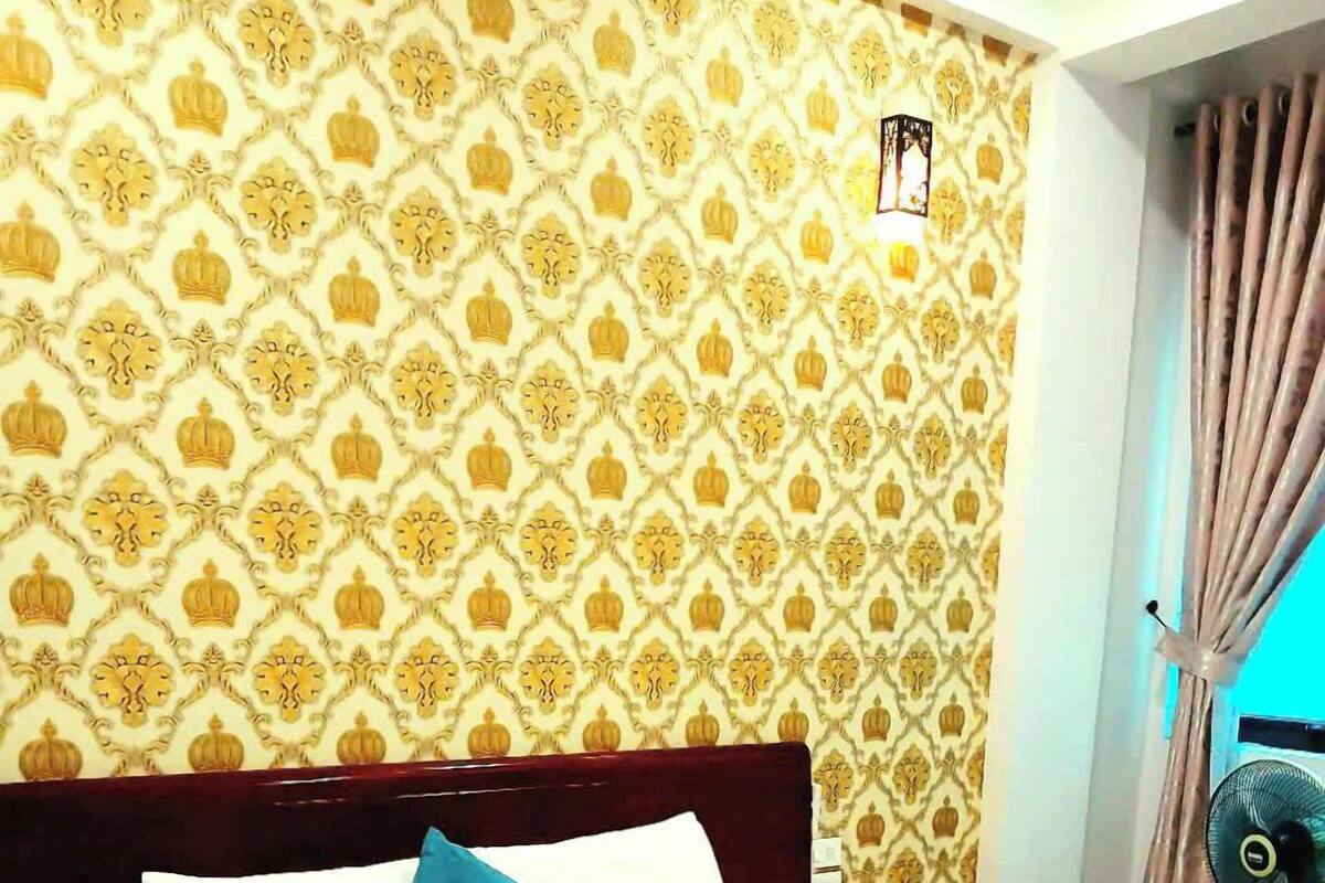 Ein Hotelzimmer mit gemustertem Wandpapier, einem Bett mit weißer Bettwäsche und einem blauen Kissen, einem Nachttisch mit Lampe sowie einem Fenster mit Vorhängen.