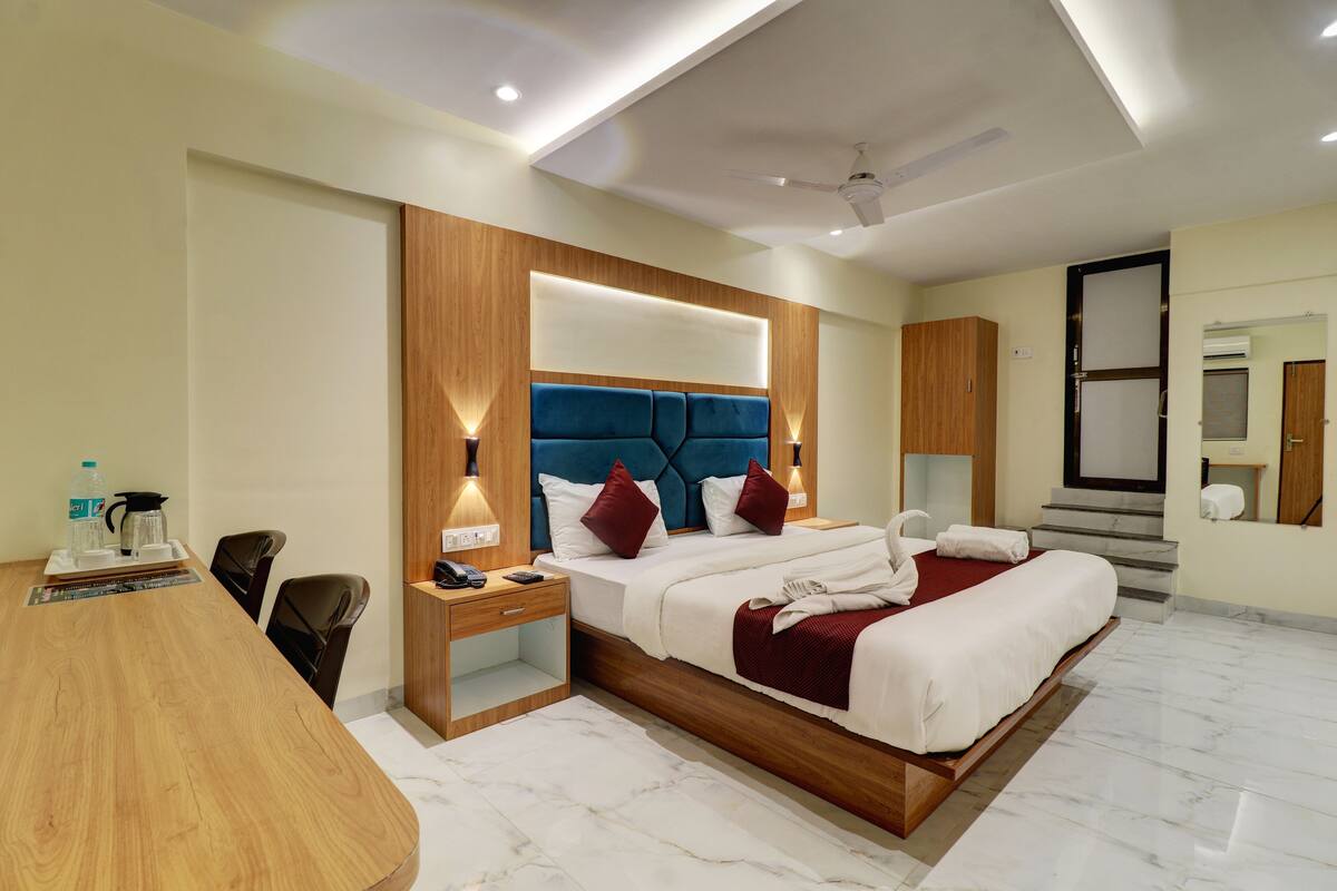 A modern hotel room with a large bed, wooden headboard, bedside lamps, a desk with a chair, and a bathroom with a shower and toilet.