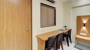 Una habitación con puerta de madera, ventana con persianas, un escritorio con sillas y una mesita de noche con lámpara.