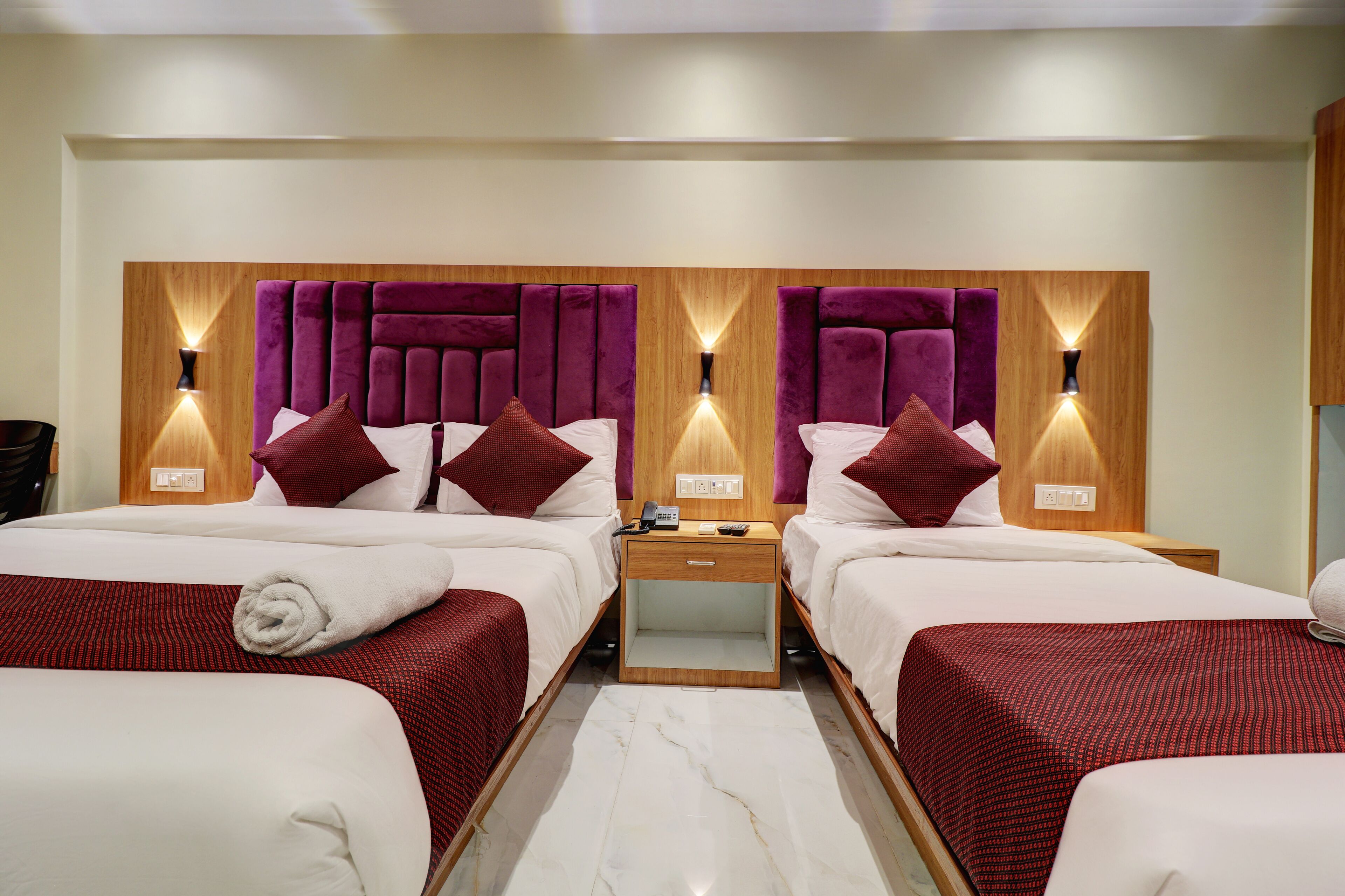 A hotel room with two beds, each with a wooden headboard, white bedding, and red accents. There are bedside tables with lamps and a small drawer.
