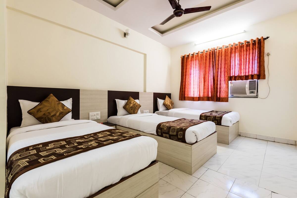 A hotel room with three beds, a ceiling fan, an air conditioning unit, and a window with orange curtains.