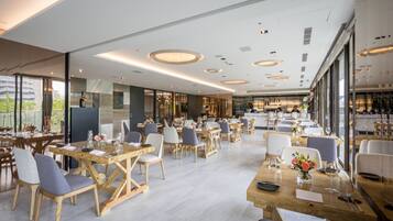 A spacious restaurant with wooden tables and chairs, large windows, and a modern ceiling design.