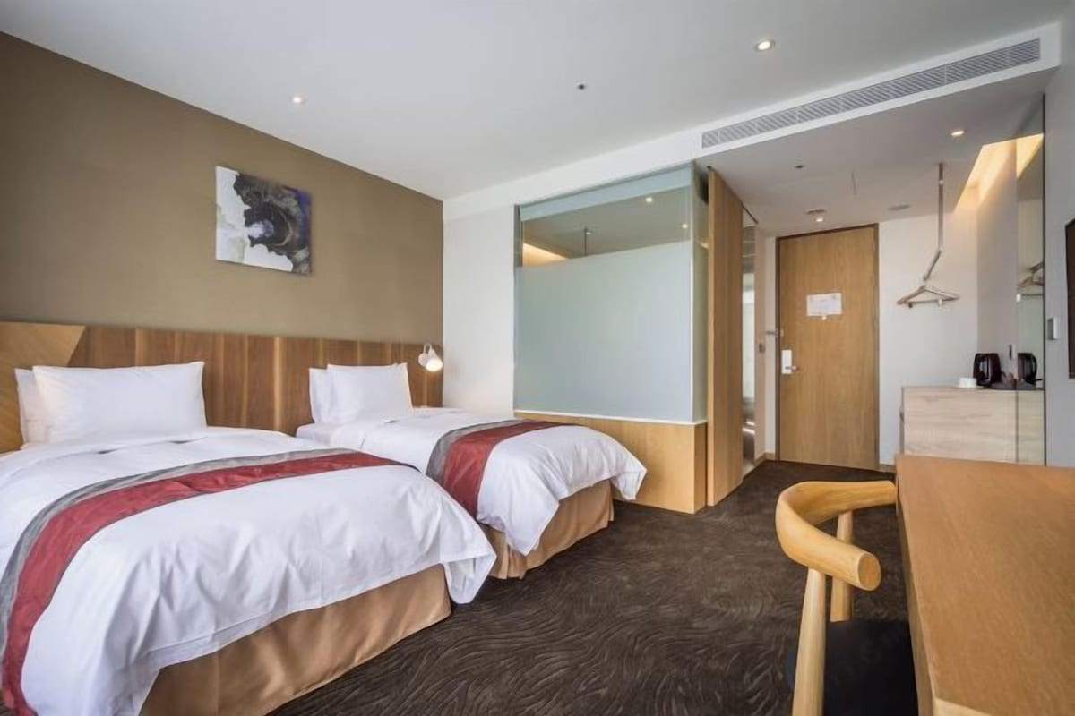 A hotel room with two beds, a desk, a chair, a coat rack, and a mirror.