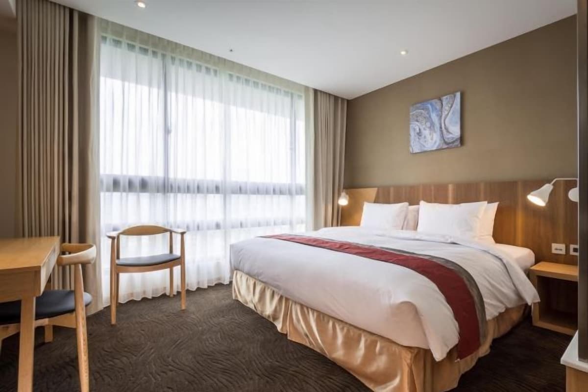 A hotel room with a bed, a desk, a chair, a large window with curtains, a wall-mounted artwork, and a nightstand with a lamp.