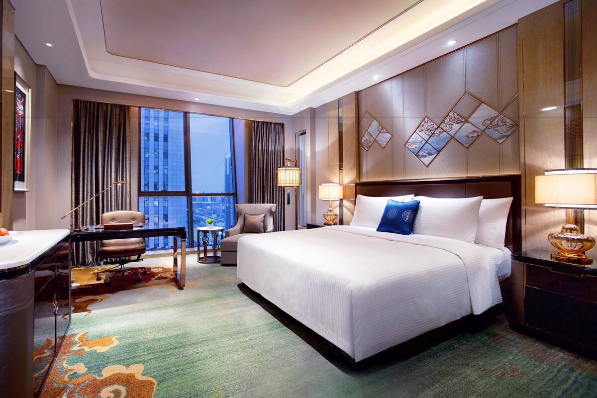 Habitación de hotel con una cama grande, un escritorio con silla, televisor y vistas de la ciudad a través de grandes ventanales.