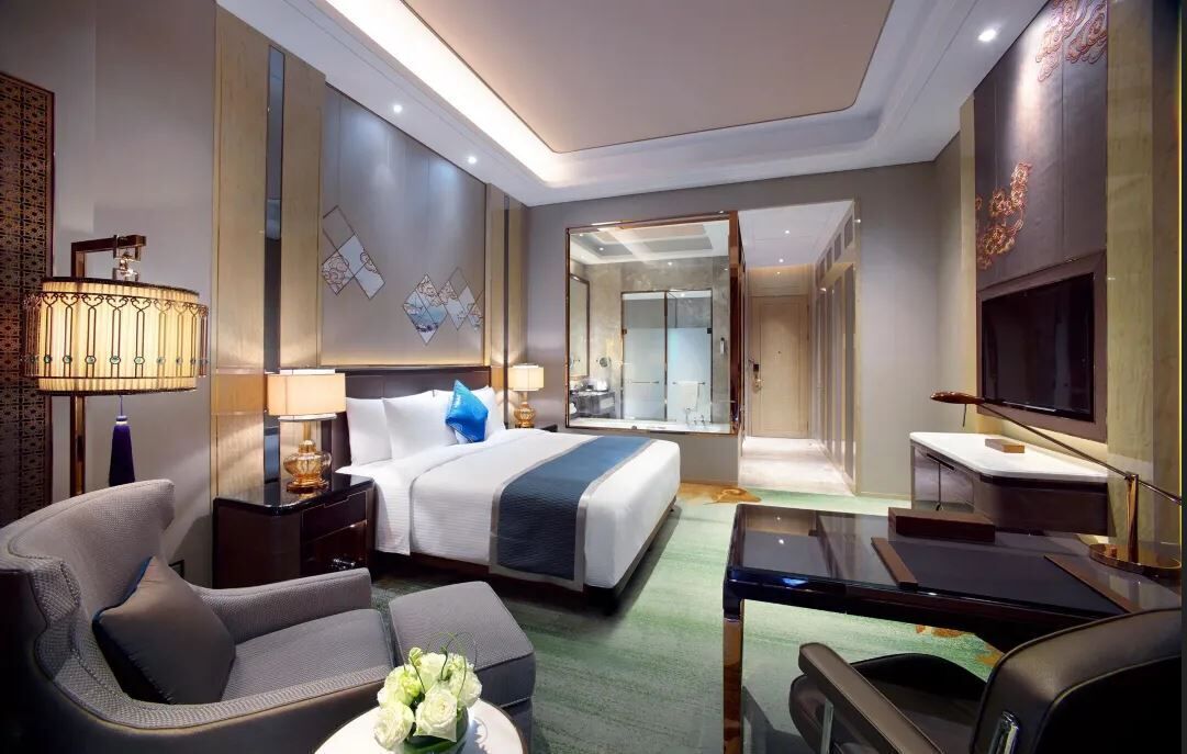 Quarto de hotel moderno com cama, escrivaninha, cadeira, televisão e banheiro visível por uma porta aberta.