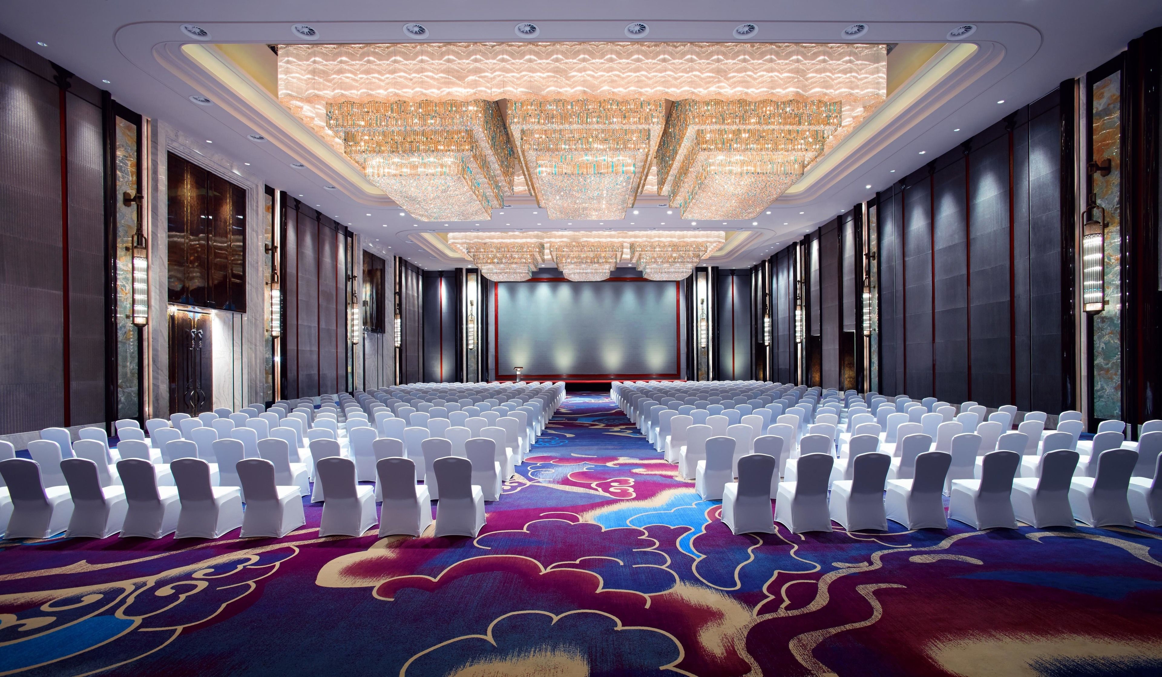 Un salón de conferencias con filas de sillas blancas, una pantalla grande al frente y una alfombra estampada.
