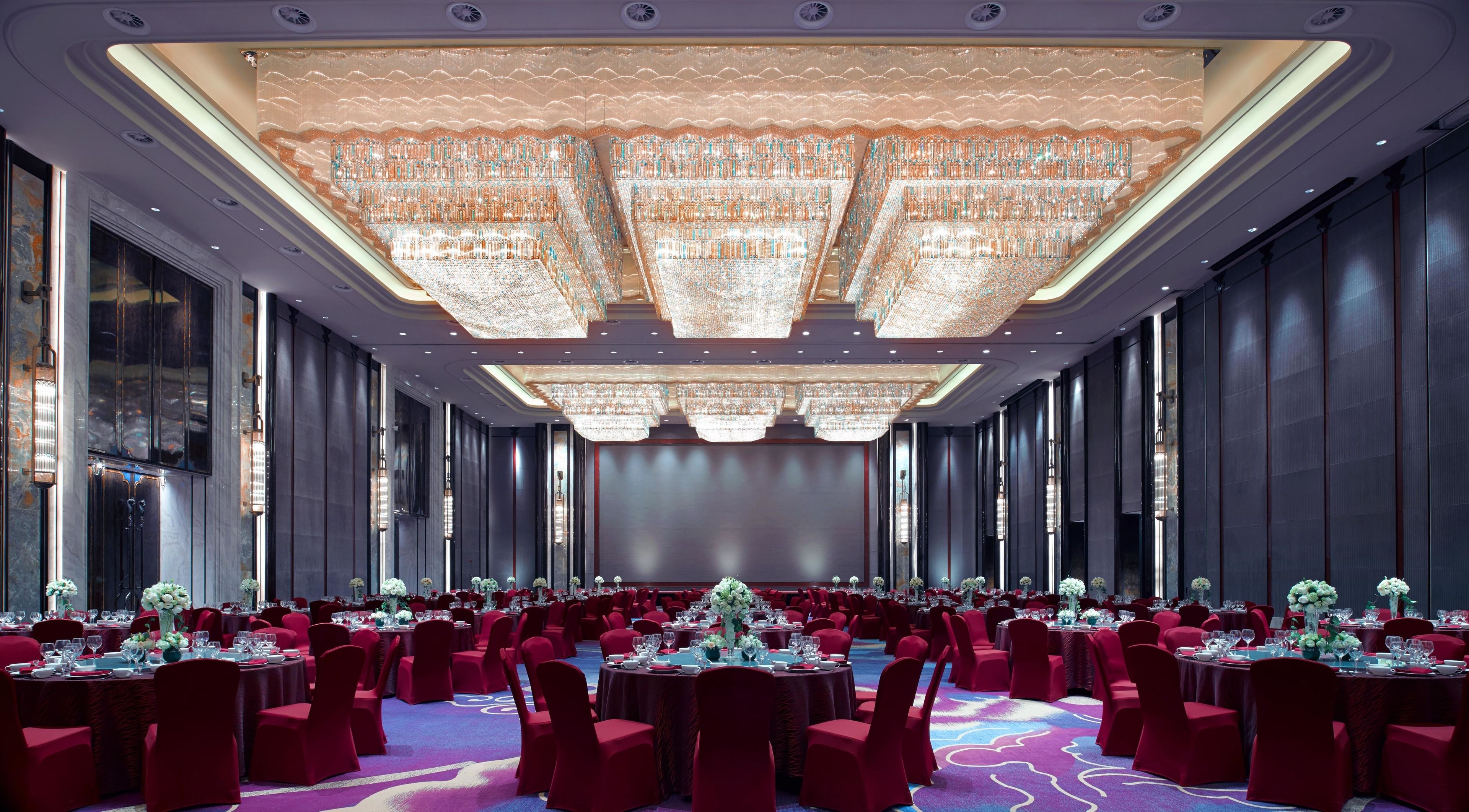 A banquet hall with round tables, red chairs, and a large chandelier.