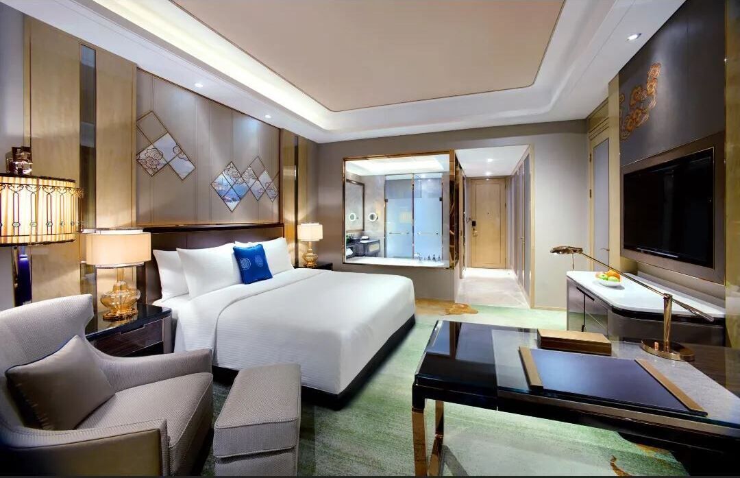Quarto de hotel com cama, escrivaninha, televisão, luminária e vista para o exterior.