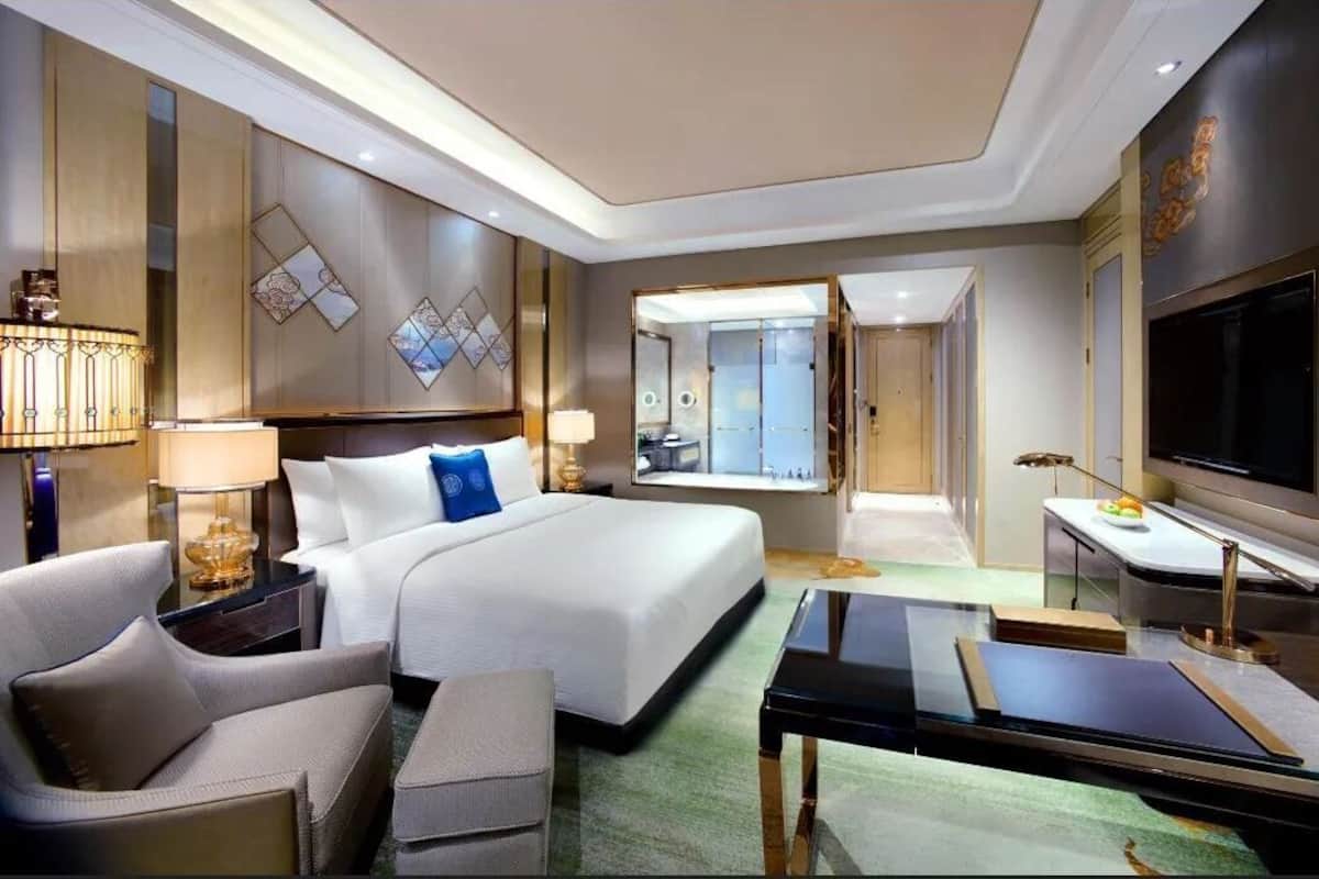 Quarto de hotel com cama, escrivaninha, televisão, luminária e vista para o exterior.