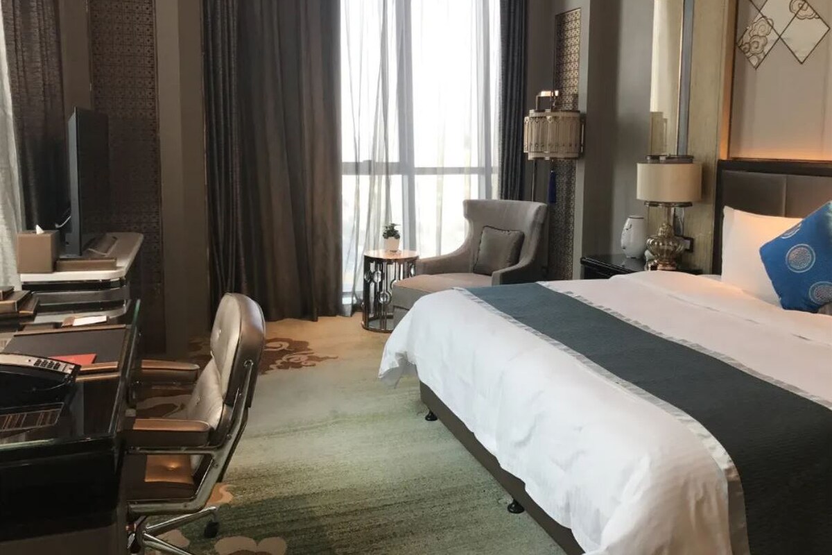 Habitación de hotel con cama, escritorio, silla, lámpara y vistas al exterior.