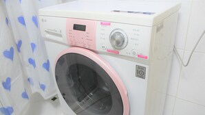 A front-loading washing machine with a digital display and control panel, located in a bathroom with white tiled walls and a shower curtain with heart patterns. - Hongstay in Hongdae (Seoul)